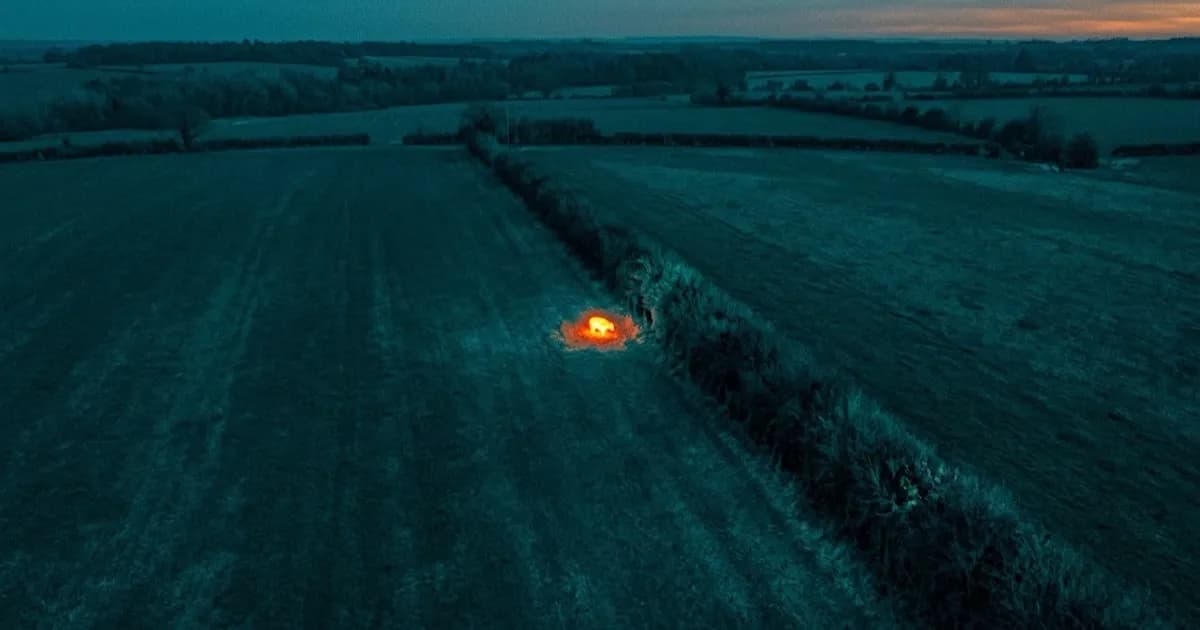 Thermal-style drone camera view over an open dark field at dusk, a bright heat signature visible at the edge of a hedgerow
